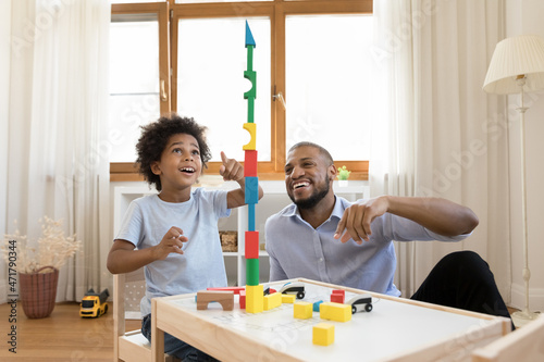 Excited Black little son and happy dad building high toy tower together, playing with construction blocks for kids, completing model, enjoying family playtime, game, entertainment at home. Fatherhood