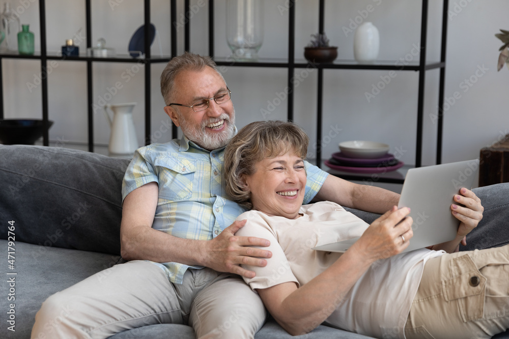 Cheerful old couple resting on comfortable couch together, using laptop, watching movie, smiling, laughing, hugging, enjoying leisure time. Happy husband and wife making video call to family Cheerful old couple resting on comfortable couch together, using laptop, watching movie, smiling, laughing, hugging, enjoying leisure time. Happy husband and wife making video call to family