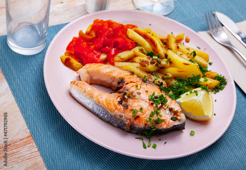 Salmon steak with fried potato, red pepper and slice of lemon served on white plate with serving pieces.