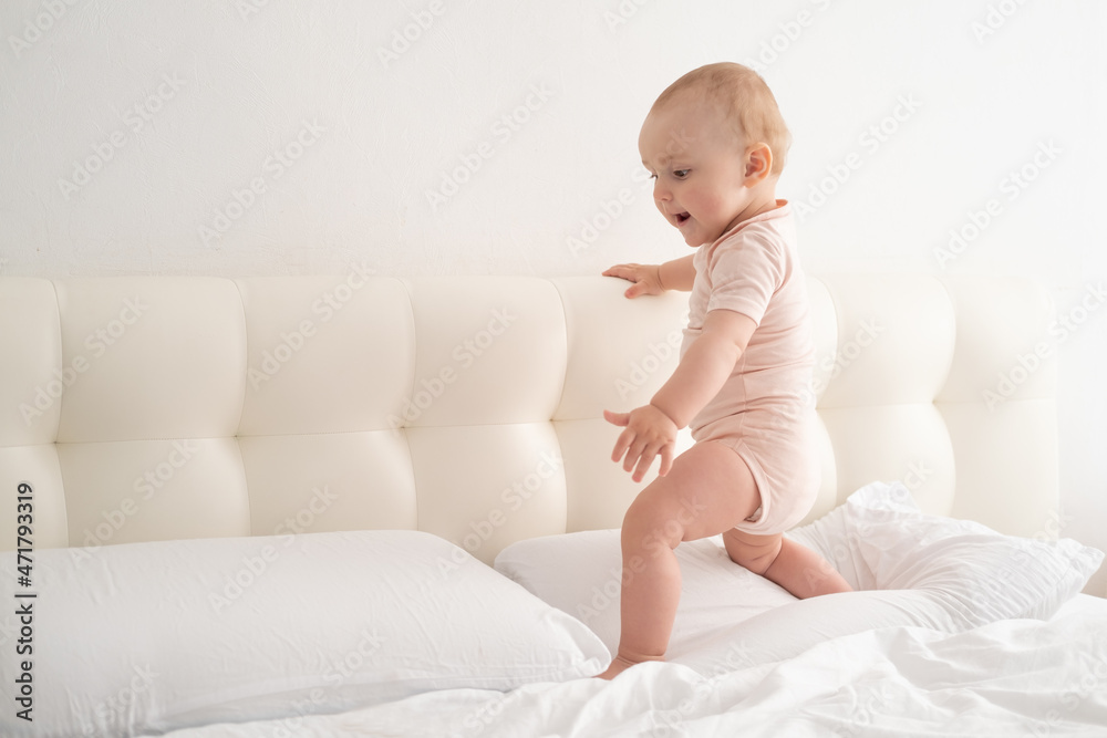 Cute baby girl in bodysuit stand holding bed frame, learning to walk on white bedding on bed