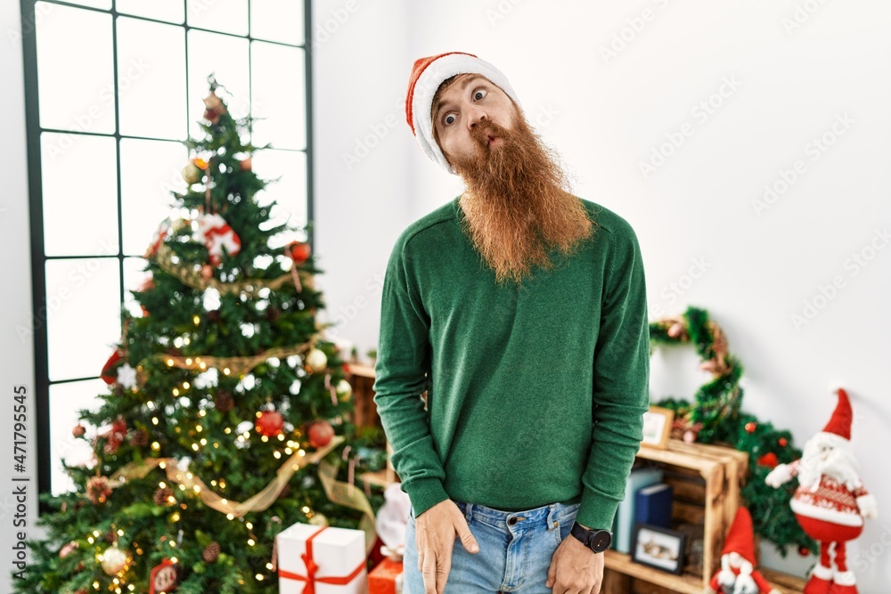 Redhead man with long beard wearing christmas hat by christmas tree making fish face with lips, crazy and comical gesture. funny expression. Redhead man with long beard wearing christmas hat by christmas tree making fish face with lips, crazy and comical gesture. funny expression.