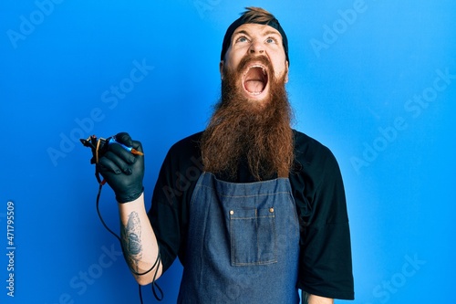 Redhead man with long beard tattoo artist wearing professional uniform and gloves angry and mad screaming frustrated and furious, shouting with anger. rage and aggressive concept.