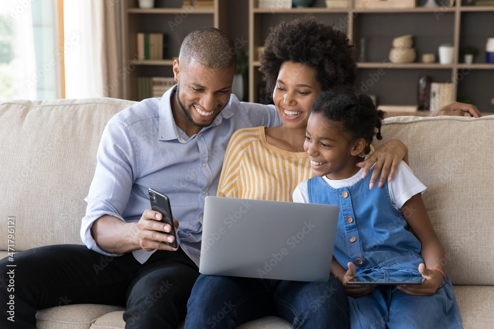 Happy African American parents and daughter kid resting on couch together, enjoying leisure with digital devices, holding, sharing laptop, smartphone, tablet, laughing, watching online content Happy African American parents and daughter kid resting on couch together, enjoying leisure with digital devices, holding, sharing laptop, smartphone, tablet, laughing, watching online content