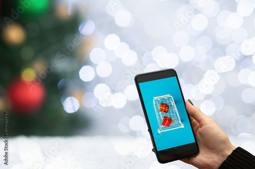 christmas shopping on internet from phone,hands with a smartphone with a cart with gifts on the background of a Christmas tree and New Year's lights