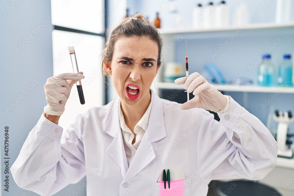 Young woman working at scientist laboratory holding blood sample clueless and confused expression. doubt concept.