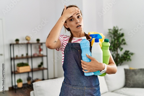 Young brunette woman wearing apron holding cleaning products stressed and frustrated with hand on head, surprised and angry face