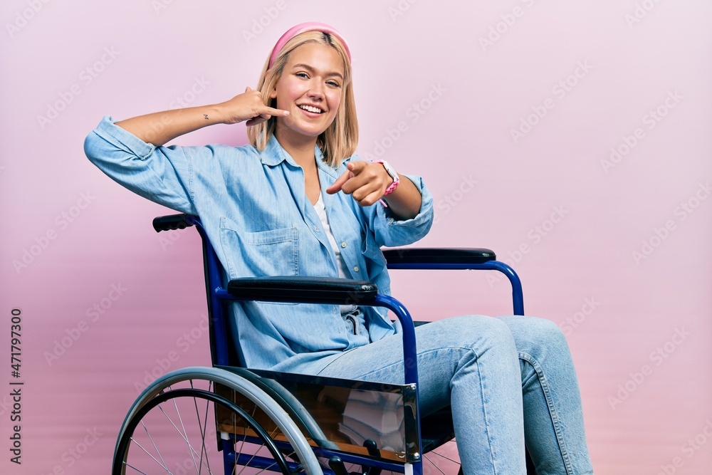Beautiful blonde woman sitting on wheelchair smiling doing talking on the telephone gesture and pointing to you. call me. Beautiful blonde woman sitting on wheelchair smiling doing talking on the telephone gesture and pointing to you. call me.