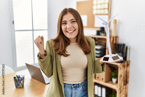 Young brunette woman holding solar windmill for renewable electricity at the office screaming proud, celebrating victory and success very excited with raised arms © Krakenimages.com