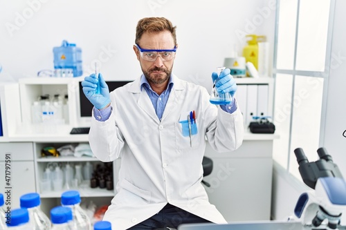 Middle age man working at scientist laboratory holding chemical products skeptic and nervous, frowning upset because of problem. negative person.