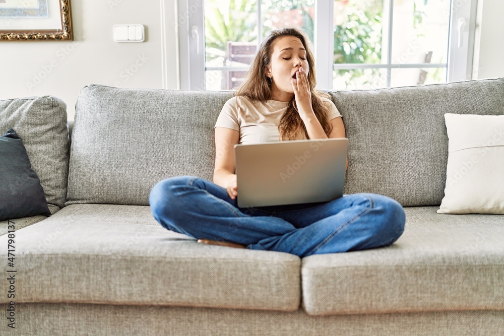 Beautiful young brunette woman sitting on the sofa using computer laptop at home bored yawning tired covering mouth with hand. restless and sleepiness. Beautiful young brunette woman sitting on the sofa using computer laptop at home bored yawning tired covering mouth with hand. restless and sleepiness.