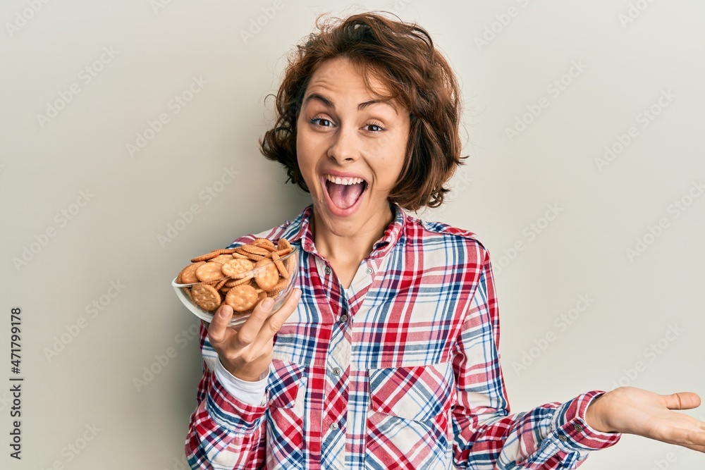 Young brunette woman holding bowl with salty biscuits celebrating achievement with happy smile and winner expression with raised hand Young brunette woman holding bowl with salty biscuits celebrating achievement with happy smile and winner expression with raised hand
