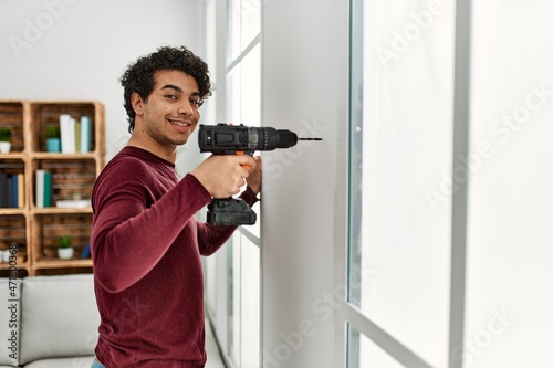 Young hispanic man smiling happy drillling wall at home.