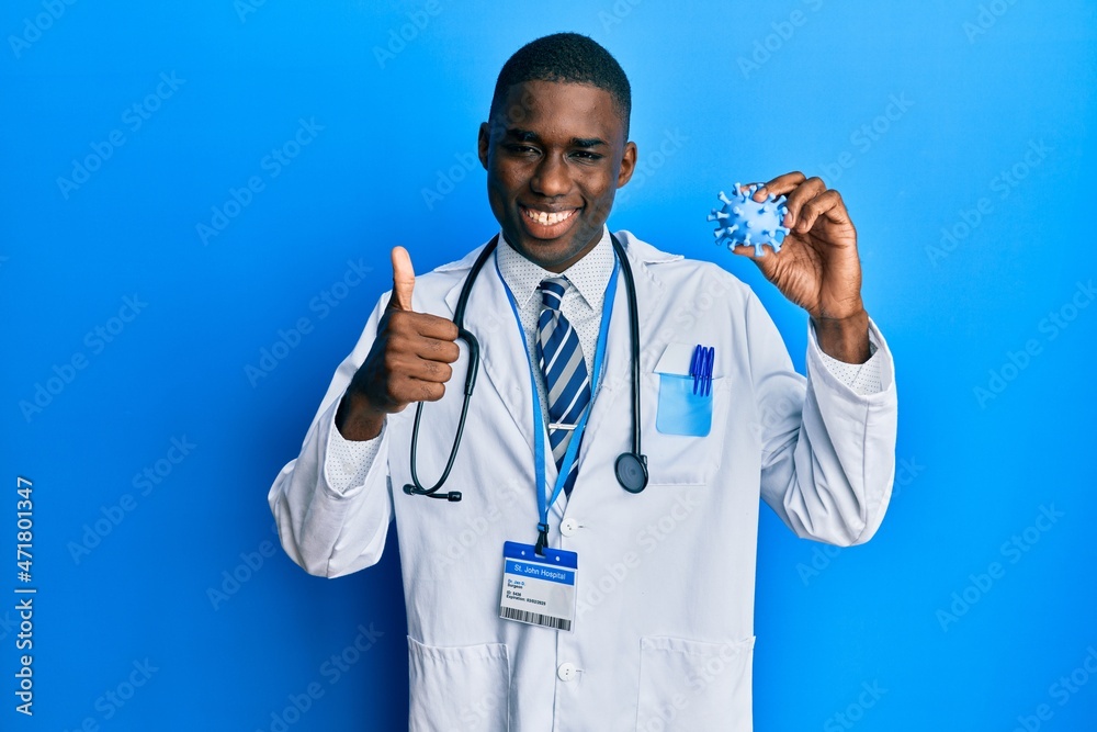 Young african american man wearing doctor coat holding virus toy smiling happy and positive, thumb up doing excellent and approval sign Young african american man wearing doctor coat holding virus toy smiling happy and positive, thumb up doing excellent and approval sign