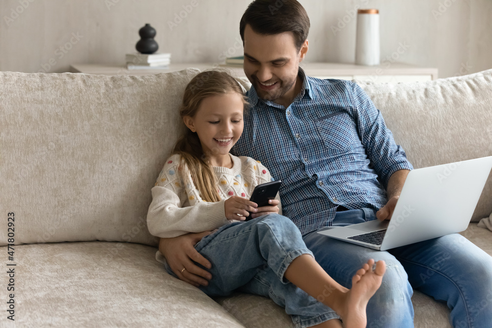 Distracted from computer work happy young father watching laughing small child daughter playing funny games on cellphone, using different gadgets, sitting on comfortable couch at home, tech addiction. Distracted from computer work happy young father watching laughing small child daughter playing funny games on cellphone, using different gadgets, sitting on comfortable couch at home, tech addiction.