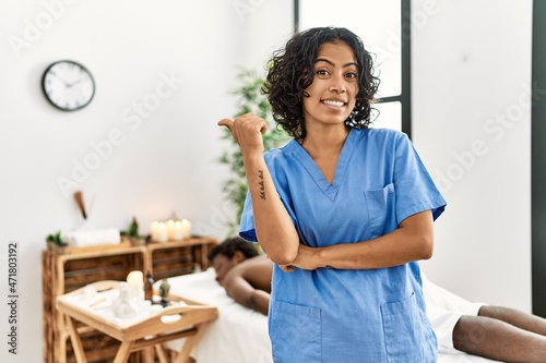 Young therapist woman at wellness spa center smiling with happy face looking and pointing to the side with thumb up.