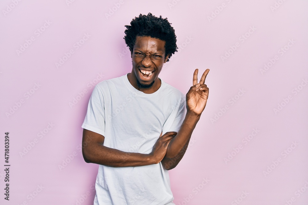 Young african american man wearing casual white t shirt smiling with happy face winking at the camera doing victory sign. number two. Young african american man wearing casual white t shirt smiling with happy face winking at the camera doing victory sign. number two.