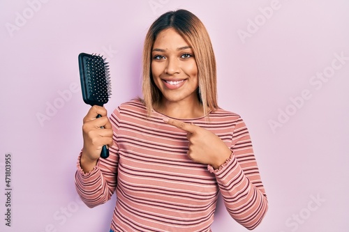 Beautiful hispanic woman holding hairbrush smiling happy pointing with hand and finger