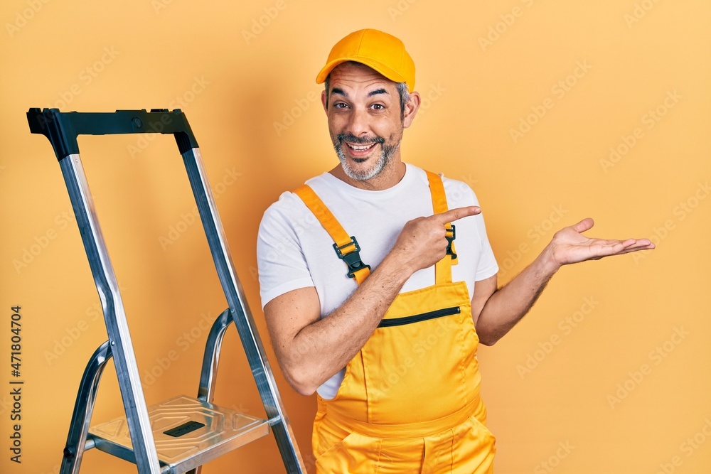 Handsome middle age man with grey hair holding ladder amazed and smiling to the camera while presenting with hand and pointing with finger.
