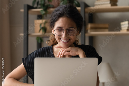 Good work. Positive smiling latina woman wear glasses sit by desk use laptop engaged in web search of information write blog online. Happy young female chat at internet read good news at social media