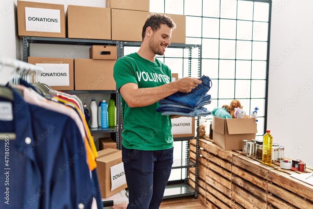 Young hispanic man wearing volunteer uniform holding folded jeans at charity center