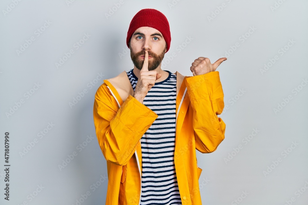 Caucasian man with beard wearing yellow raincoat asking to be quiet with finger on lips pointing with hand to the side. silence and secret concept.