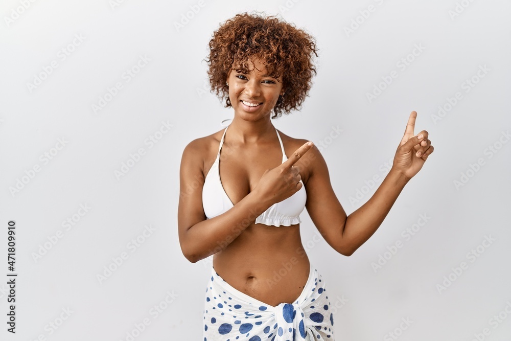 Young african american woman with curly hair wearing bikini smiling and looking at the camera pointing with two hands and fingers to the side. Young african american woman with curly hair wearing bikini smiling and looking at the camera pointing with two hands and fingers to the side.