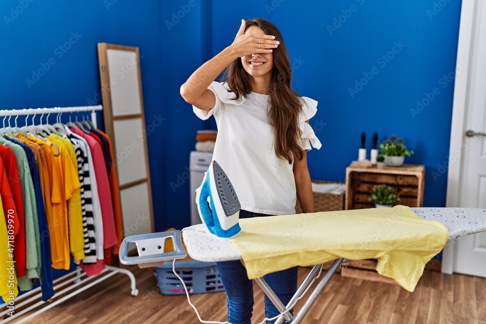 Young hispanic woman ironing clothes at laundry room smiling and laughing with hand on face covering eyes for surprise. blind concept. Young hispanic woman ironing clothes at laundry room smiling and laughing with hand on face covering eyes for surprise. blind concept.