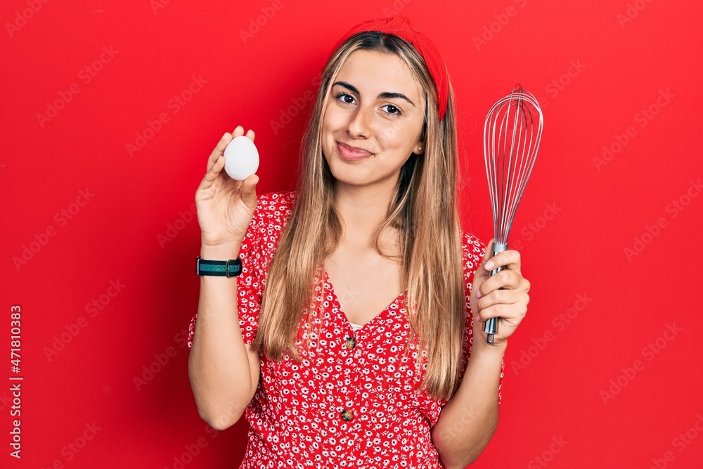 Beautiful hispanic woman holding egg and baker whisk relaxed with serious expression on face. simple and natural looking at the camera. Beautiful hispanic woman holding egg and baker whisk relaxed with serious expression on face. simple and natural looking at the camera.
