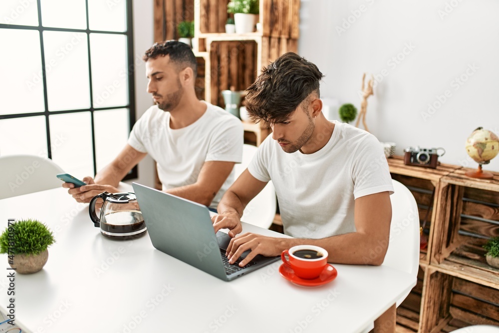 Two hispanic men couple using smartphone and laptop at home