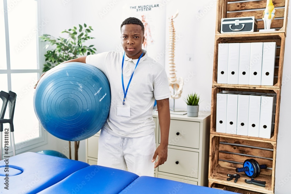 Fototapeta premium Young african physiotherapy man holding pilates ball at rehabilitation clinic thinking attitude and sober expression looking self confident