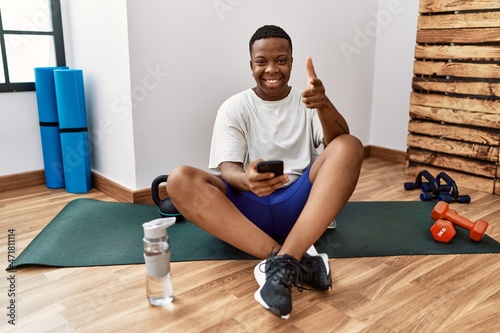 Young african man sitting on training mat at the gym using smartphone pointing fingers to camera with happy and funny face. good energy and vibes.