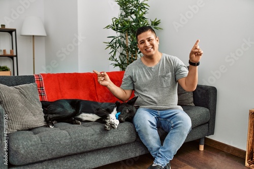 Young latin man and dog sitting on the sofa at home smiling confident pointing with fingers to different directions. copy space for advertisement © Krakenimages.com