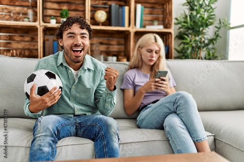 Man smiling happy watching soccer match and girlfriend boring using smartphone at home.