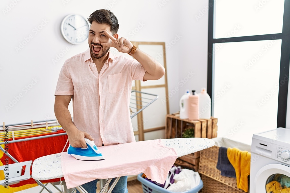 Young man with beard ironing clothes at home doing peace symbol with fingers over face, smiling cheerful showing victory Young man with beard ironing clothes at home doing peace symbol with fingers over face, smiling cheerful showing victory