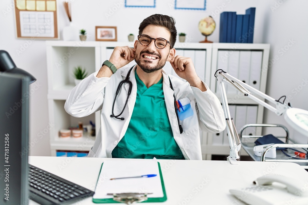 Young man with beard wearing doctor uniform and stethoscope at the clinic smiling pulling ears with fingers, funny gesture. audition problem Young man with beard wearing doctor uniform and stethoscope at the clinic smiling pulling ears with fingers, funny gesture. audition problem