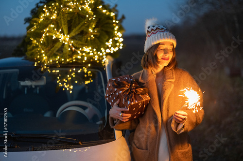 Happy caucasian woman firing sparklers near car with Christmas tree on nature at dusk. Concept of celebrating New Year holidays alone. Idea of Christmas mood and fun
