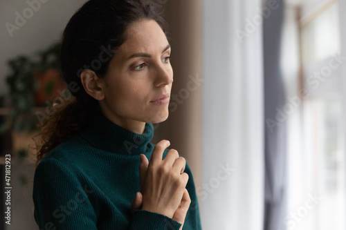 Unhappy thoughts. Worried young latin woman look at window feel lonely sad wait for boyfriend husband coming back after quarrel. Pessimistic millennial lady hide from life problems at home. Copy space photo
