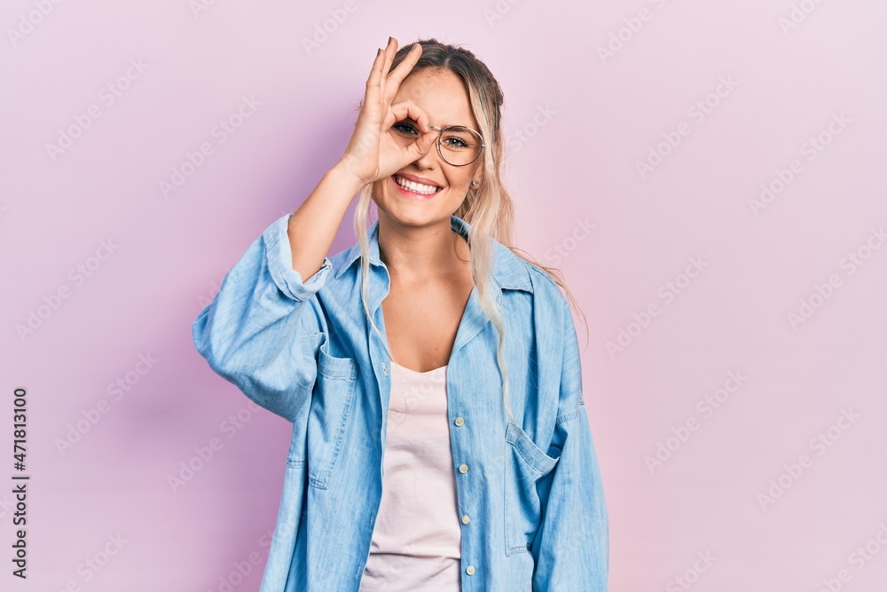 Beautiful young blonde woman wearing casual clothes and glasses doing ok gesture with hand smiling, eye looking through fingers with happy face.