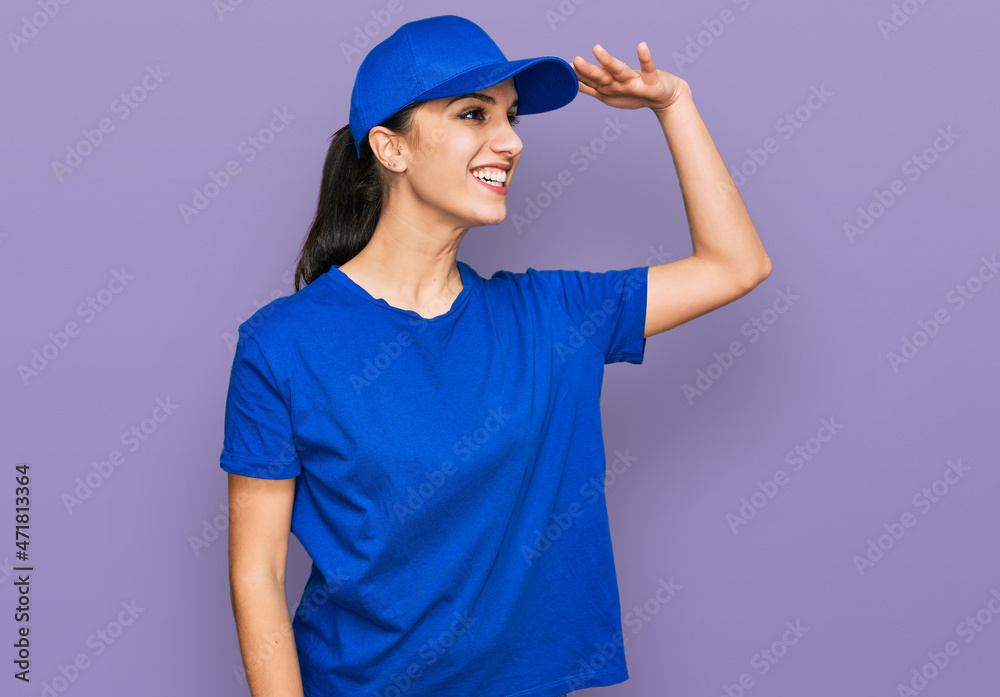 Young hispanic girl wearing delivery courier uniform very happy and smiling looking far away with hand over head. searching concept. Young hispanic girl wearing delivery courier uniform very happy and smiling looking far away with hand over head. searching concept.