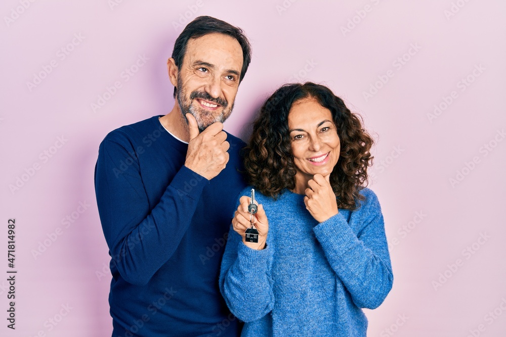 Middle age couple of hispanic woman and man holding keys of new home looking confident at the camera smiling with crossed arms and hand raised on chin. thinking positive.