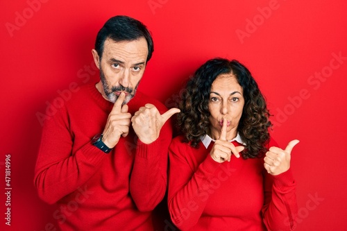 Middle age couple of hispanic woman and man hugging and standing together asking to be quiet with finger on lips pointing with hand to the side. silence and secret concept.