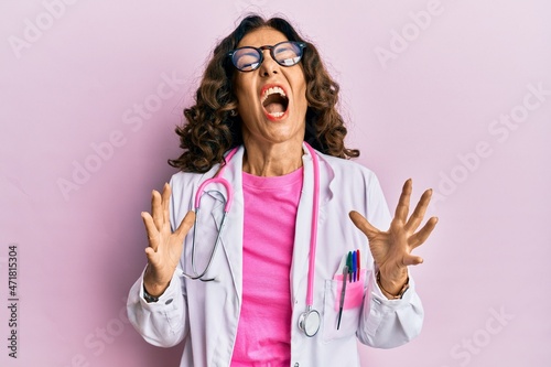 Middle age hispanic woman wearing doctor uniform and glasses crazy and mad shouting and yelling with aggressive expression and arms raised. frustration concept.