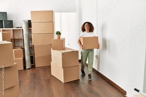 Middle age hispanic woman smiling confident holding cardboard box at new home