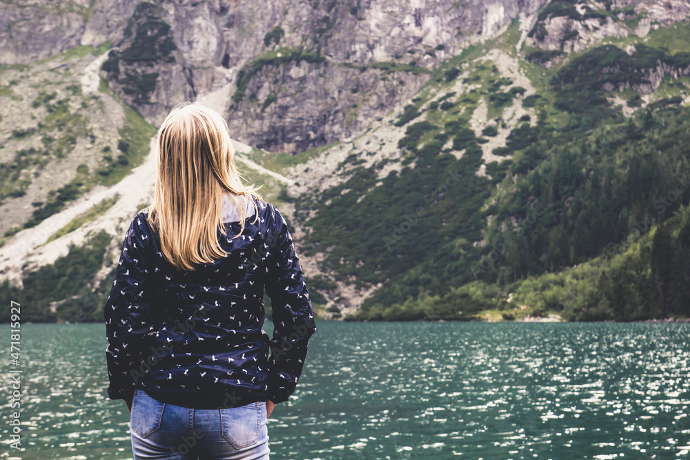 A young blonde girl stands back and looks at blue mountain lake and the mountains A young blonde girl stands back and looks at blue mountain lake and the mountains
