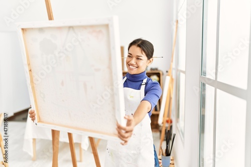 Young latin woman smiling confident holding canvas draw at art studio