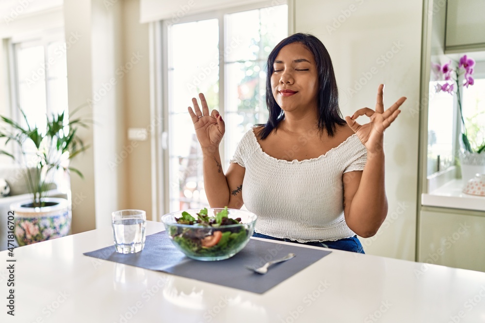 Young hispanic woman eating healthy salad at home relax and smiling with eyes closed doing meditation gesture with fingers. yoga concept. Young hispanic woman eating healthy salad at home relax and smiling with eyes closed doing meditation gesture with fingers. yoga concept.