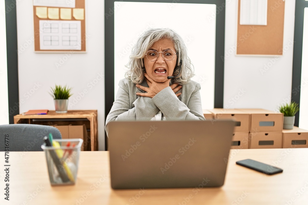 Middle age businesswoman sitting on desk working using laptop at office shouting and suffocate because painful strangle. health problem. asphyxiate and suicide concept. Middle age businesswoman sitting on desk working using laptop at office shouting and suffocate because painful strangle. health problem. asphyxiate and suicide concept.