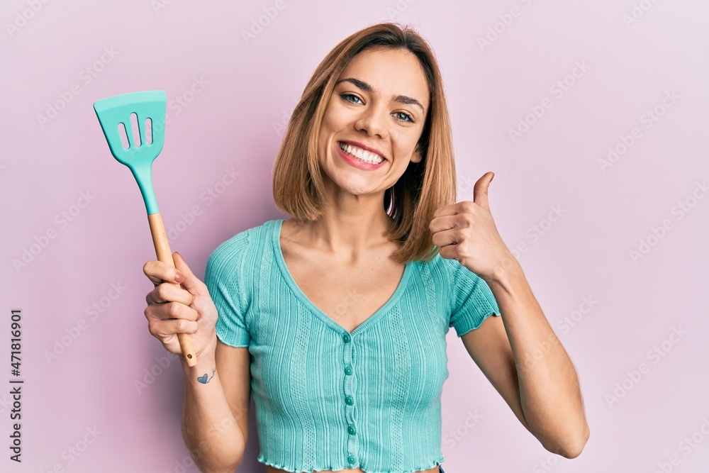 Young caucasian blonde woman holding silicone spatula smiling happy and positive, thumb up doing excellent and approval sign Young caucasian blonde woman holding silicone spatula smiling happy and positive, thumb up doing excellent and approval sign