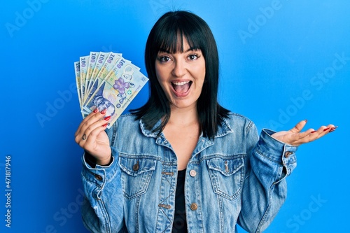 Young hispanic woman holding 100 romanian leu banknotes celebrating achievement with happy smile and winner expression with raised hand photo