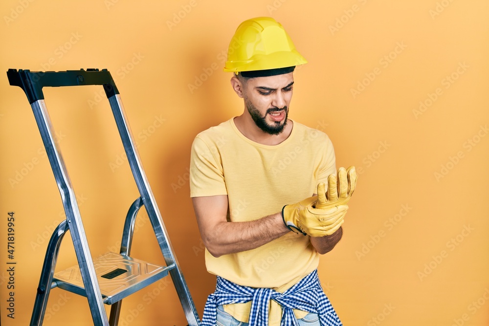 Handsome man with beard by construction stairs wearing hardhat suffering pain on hands and fingers, arthritis inflammation Handsome man with beard by construction stairs wearing hardhat suffering pain on hands and fingers, arthritis inflammation